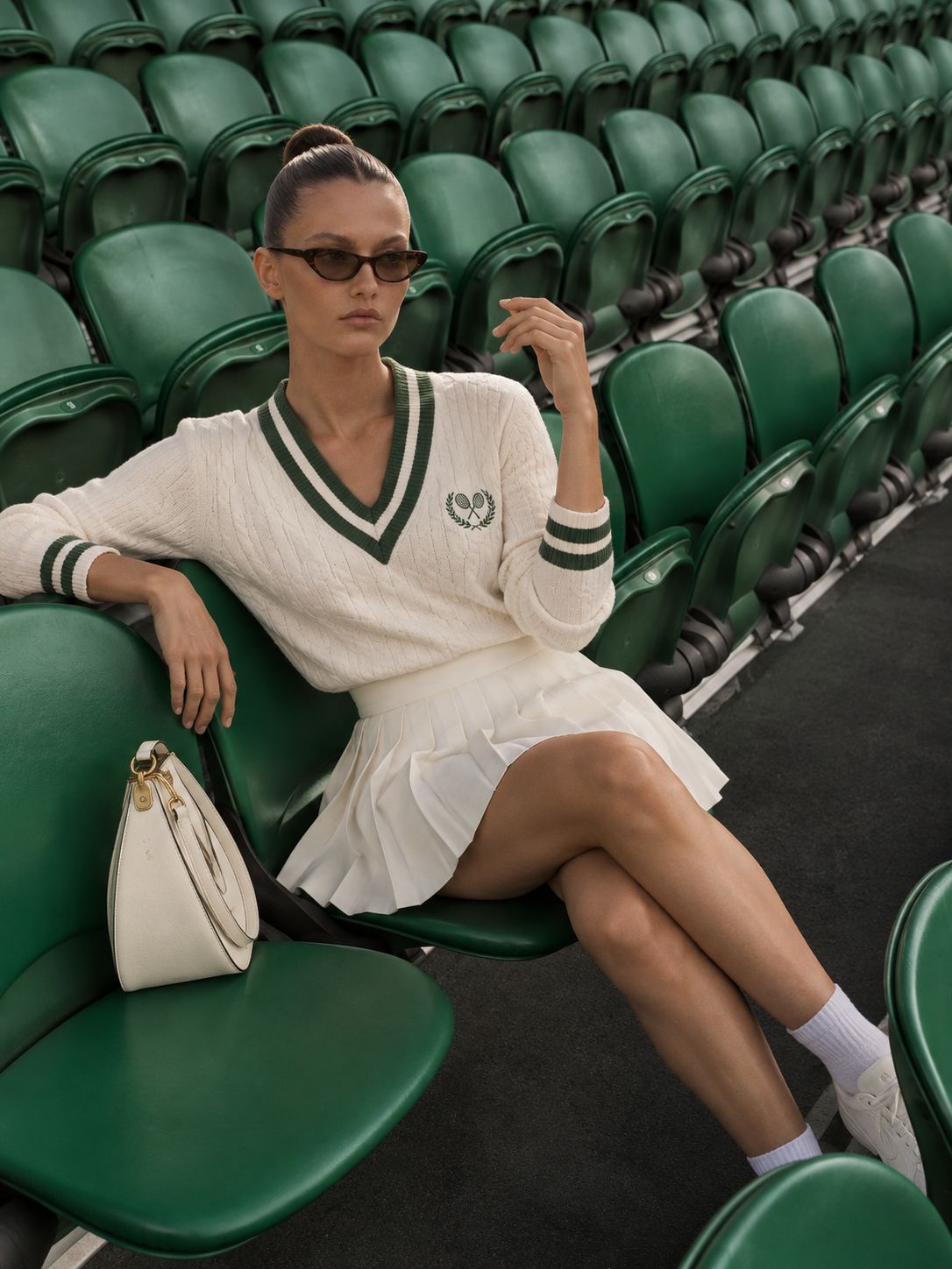 A woman in a cream cable-knit tennis sweater and pleated skirt sitting in dark green stadium seats.